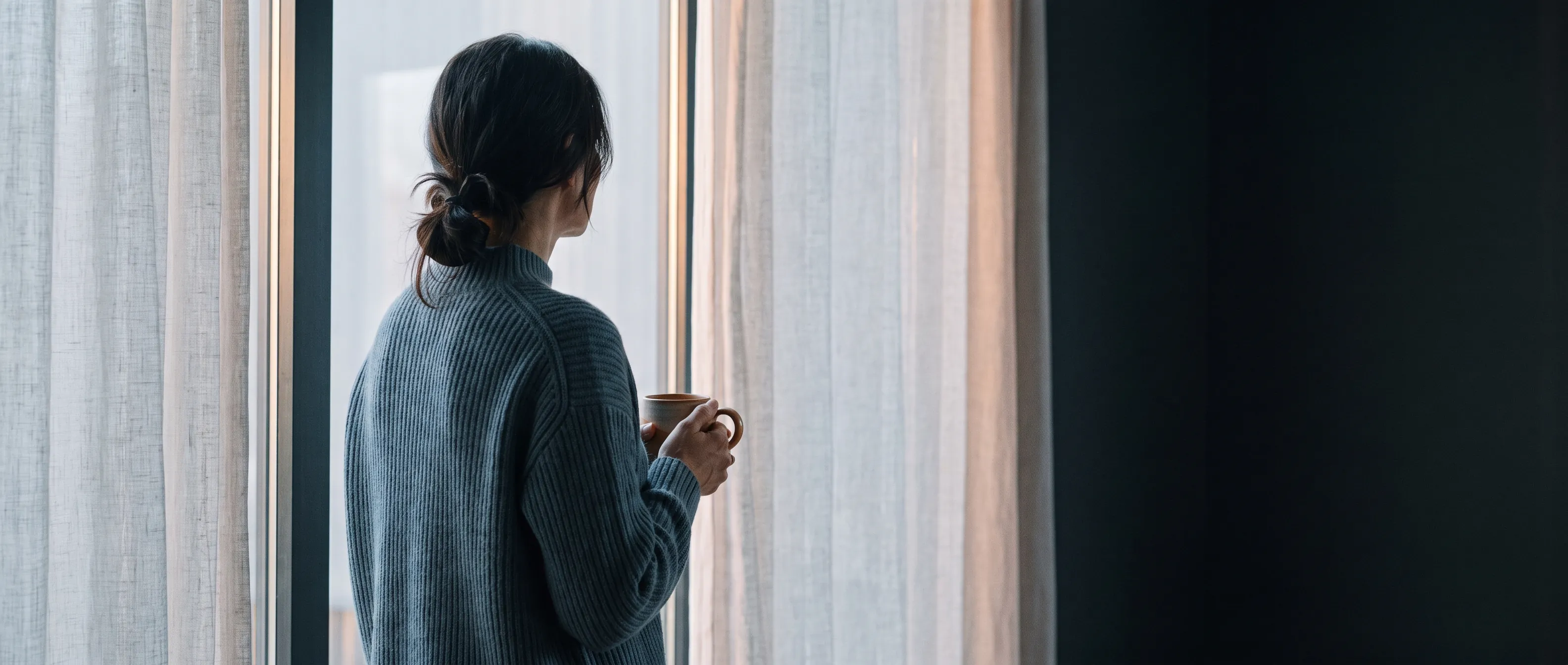 A woman holding a mug, looking out a bright window in soft morning light